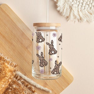 May include: A clear glass reusable water bottle with a bamboo lid and a straw. The bottle is decorated with a pattern of leopard print bunnies and pink flowers.