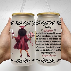 May include: Two frosted glass jars with wooden lids. The jars have a black and white leopard print design. One jar has a graduation cap and gown illustration of a Black woman holding a diploma and flowers. The other jar has text that reads "To My Daughter You believed you could, so you did. You have brains in your head. You have feet in your shoes. You can steer yourself in any direction you choose. Take pride in how far you've come. Have faith in how far you can go. But don't forget to enjoy the journey. Congratulations!"