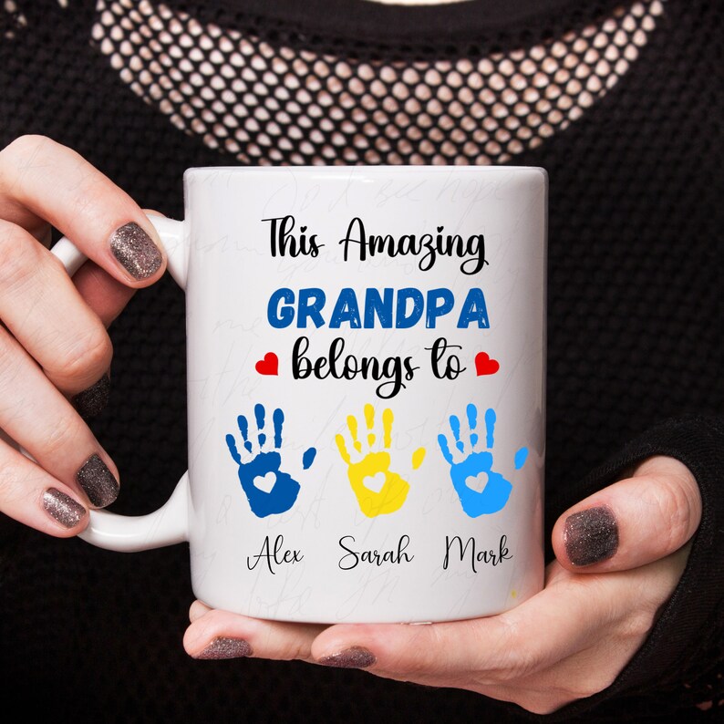 May include: White ceramic mug with the text "This Amazing Grandpa belongs to" and three handprints with the names Alex, Sarah, and Mark underneath.
