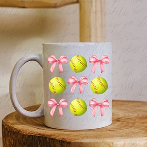 May include: White ceramic mug with a pink bow and yellow softball design. The mug has a handle and is decorated with multiple softball and bow designs.