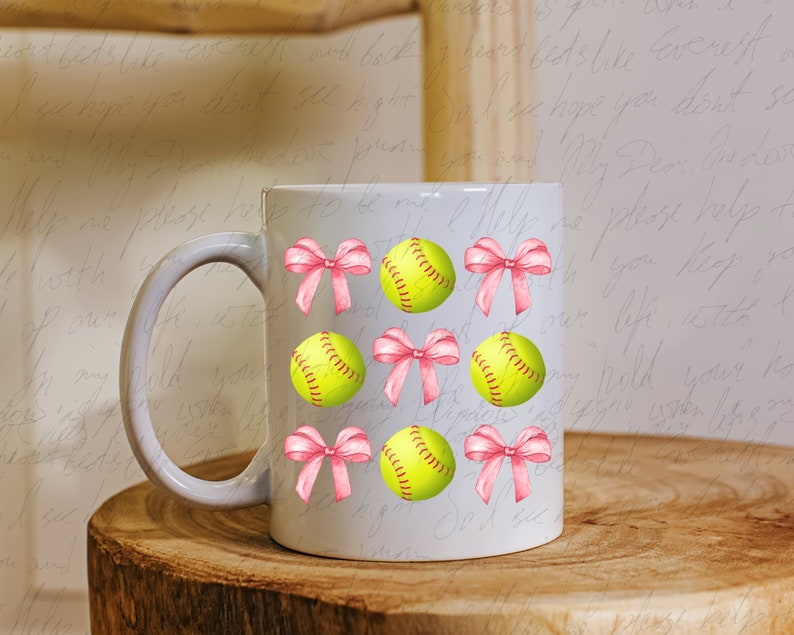 May include: White ceramic mug with a pink bow and yellow softball design. The mug has a handle and is decorated with multiple softball and bow designs.