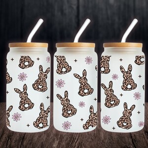 May include: Three glass tumblers with bamboo lids and straws. Each tumbler features a white background with a repeating pattern of pink flowers and brown leopard print bunnies.