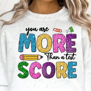 May include: White sweatshirt with the text "you are MORE than a test SCORE" in colorful glitter letters. The design includes a pencil, paperclip, and push pin. The sweatshirt is a comfortable, casual style.