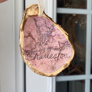May include: Gold oyster shell ornament with a pink map of Charleston, South Carolina inside. The ornament has a white ribbon for hanging.