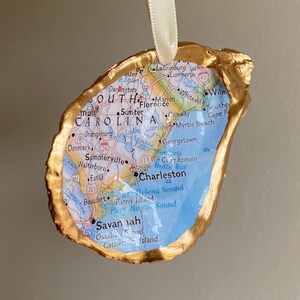 May include: A gold-painted oyster shell ornament with a map of South Carolina inside. The map shows major cities and towns, including Charleston, Savannah, and Myrtle Beach.