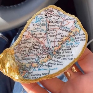 May include: A gold-painted oyster shell with a map of Charleston, South Carolina inside. The map shows the city's major landmarks, including the Charleston Harbor, Fort Sumter, and Sullivan's Island.