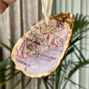 May include: Gold-painted oyster shell ornament with a vintage map of Charleston, South Carolina, featuring the text "Charleston, Ft. Sumter, Beaufort, Edisto, Rice, Phosphates".
