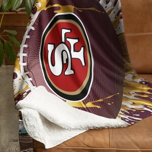 May include: A burgundy and gold football-themed throw blanket featuring the San Francisco 49ers logo. The blanket has a white sherpa lining and is draped over a brown leather couch. The design includes a football graphic with gold paint splatter.