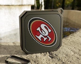 May include: A black, square-shaped portable speaker with a red and gold logo. The logo features the letters "SF" in white. A black carabiner clip rests below the speaker on a stone surface. The speaker has a loop for hanging.