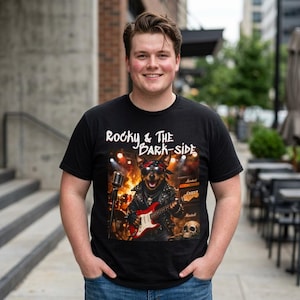May include: Black t-shirt featuring a dog playing a red electric guitar on stage. The shirt has the text "Rocky & The Bark-Side" above the dog graphic. The dog is wearing a bandana and a leather jacket.