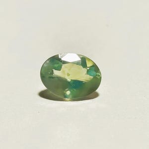 May include: An oval-shaped gemstone with a blend of green and yellow tones. The gem features a faceted surface that reflects light, producing a sparkling effect. The stone is set against a plain white background, accentuating its colour and shape.