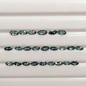 Puede incluir: Una variedad de gemas de talla marquesa de color verde azulado, dispuestas en filas sobre una superficie blanca. Las gemas, de forma ovalada con extremos puntiagudos, muestran tonos que van del verde azulado claro al oscuro. La disposición resalta la forma y el color de las gemas.