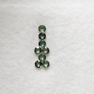 Puede incluir: Una colección de ocho gemas redondas y facetadas en varios tonos de verde, dispuestas en una línea vertical. Las gemas se muestran sobre un fondo blanco texturizado, mostrando su claridad y color. Adecuado para la fabricación de joyas.