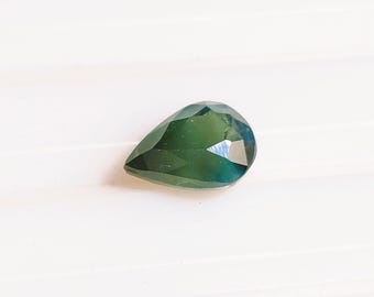 May include: A pear-shaped loose gemstone featuring a deep forest green hue with subtle blue undertones. The stone has a faceted surface that reflects light, creating a polished appearance with sharp edges and a pointed top section.