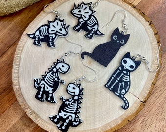 Puede incluir: Un conjunto de cinco pendientes de esqueleto con un gato negro, un dinosaurio y tres tipos diferentes de dinosaurios. Los pendientes están hechos de acrílico blanco y tienen ganchos plateados.