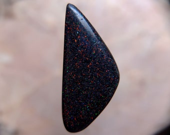 May include: A black opal cabochon with a triangular shape and a dark, iridescent surface. The opal displays flashes of red, green, and blue.