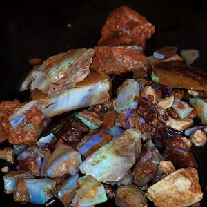 May include: A collection of raw opal stones featuring a mix of earthy brown, tan, and rust-colored rock matrix embedded with iridescent flashes of blue, green, and violet light. The stones vary in size and shape, displaying natural textures.