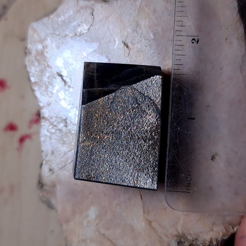 Może przedstawiać: Czarny, tęczowy kamień szlachetny o szorstkiej, fakturowanej powierzchni. Kamień szlachetny ma około 2,5 cm.