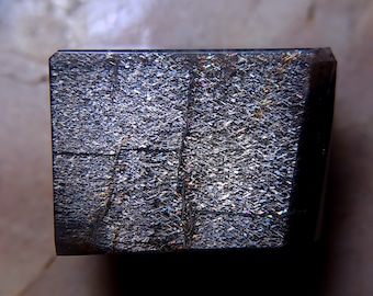 May include: A dark gray iridescent crystal with a cubic shape. The crystal has a rough, textured surface and reflects light in a rainbow of colors.