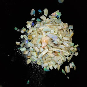 May include: A pile of white, iridescent opal chips and nuggets on a black background. The opal chips are various sizes and shapes, with some showing flashes of color.