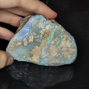 May include: A raw, unpolished opal specimen featuring a translucent, pale blue body color with patches of earthy, beige-colored matrix and vibrant flashes of green and blue iridescence visible across the uneven, textured surface of the stone.