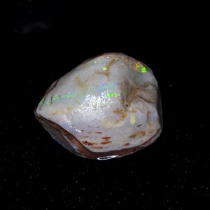 May include: A polished opal gemstone with a white base color and iridescent flashes of green, blue, and yellow. The stone has a rounded shape with a brown edge, set against a black background.