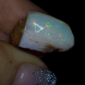 May include: A rough opal gemstone with a light blue base colour and iridescent flashes of green, yellow, and red. The stone has a rough, natural texture and a brown matrix. The opal is held in a hand.