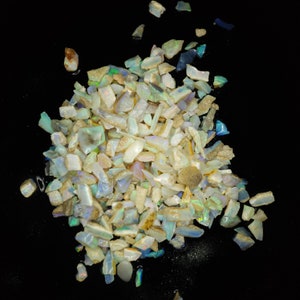 May include: A pile of small, iridescent opal chips in various shades of white, blue, and green. The chips are scattered on a black background.