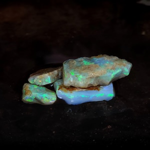 May include: A collection of raw opal stones featuring a milky white base with vibrant flashes of neon green and blue play-of-color. The stones have irregular, jagged edges and a natural, unpolished texture, showcasing internal light reflections.