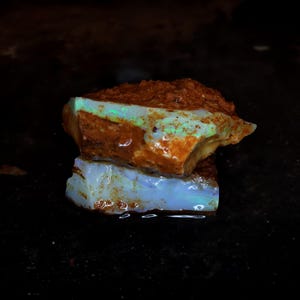 May include: A raw, unpolished opal specimen featuring a rough, earthy brown exterior with patches of iridescent green and blue play-of-color. The stone displays a translucent, milky white base with visible mineral inclusions and a jagged texture.