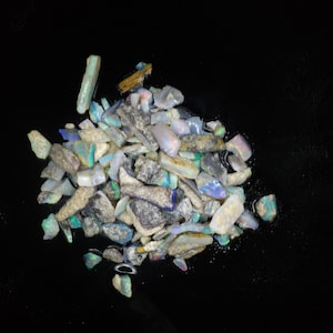 Puede incluir: Un montón de piedras preciosas de ópalo en bruto, iridiscentes, de diversas formas y tamaños. Los ópalos son una mezcla de blanco, gris y azul con destellos de colores arcoíris.