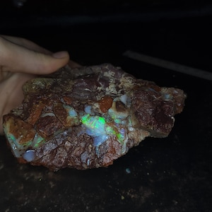 May include: A raw, unpolished rock specimen featuring a dark reddish-brown matrix with embedded patches of iridescent opal. The opal displays vibrant flashes of neon green and blue light against the textured, earthy surface of the stone.