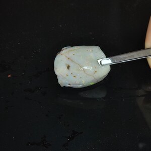 May include: A raw opal gemstone, predominantly white with hints of green and yellow iridescence. The stone features brown veining and is held by tweezers. The background is dark.