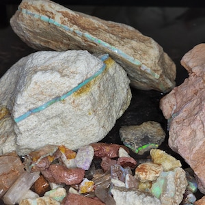 May include: Close-up of rough opal rocks in various sizes and colors. The rocks are beige, brown, and pink, with vibrant blue and green opal veins running through them. Smaller opal fragments are scattered around the larger rocks.