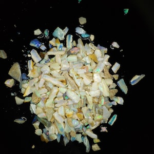May include: A pile of iridescent white opal chips on a black background. The chips are various sizes and shapes, with some showing flashes of color.