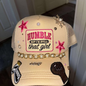 Peut inclure: Casquette de camionneur beige avec un dos en filet, ornée d'étoiles roses, de perles et d'une chaîne dorée. Le devant affiche le texte "HUMBLE but I'M STILL that girl" en rose et blanc, ainsi qu'un poing levé noir et un écusson "Queen".