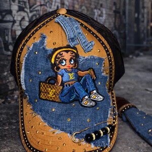 May include: A custom baseball cap featuring a denim design with a cartoon character patch. The cap has a brown brim with gold studs, a black mesh back, and a denim patch with a cartoon character. A gold safety pin and black beads are attached.