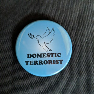 May include: A round, light blue button with the words "DOMESTIC TERRORIST" in black, beneath a white dove carrying an olive branch. The button is set against a dark, textured background.
