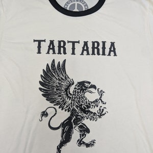 May include: A white t-shirt with a black ringer neck and a black graphic of a griffin with the word "TARTARIA" printed above it. The shirt also has a black peace sign with the words "LOVE LIFE" printed inside the peace sign.