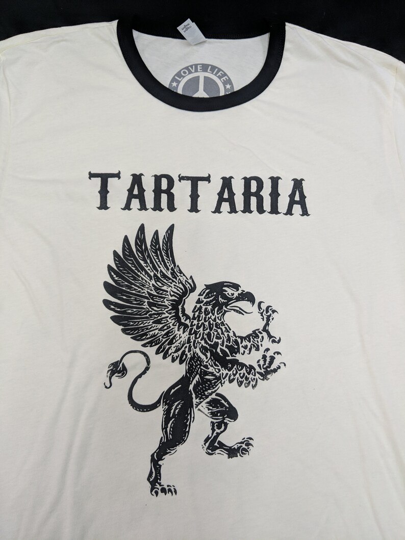 May include: A white t-shirt with a black ringer neck and a black graphic of a griffin with the word "TARTARIA" printed above it. The shirt also has a black peace sign with the words "LOVE LIFE" printed inside the peace sign.