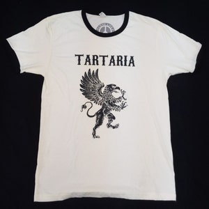 May include: White t-shirt with a black ringer and a black graphic of a griffin with the word "TARTARIA" above it.