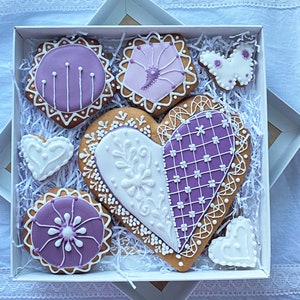 May include: A gift box filled with purple and white decorated gingerbread cookies in the shape of hearts, a butterfly, and a circle. The cookies are arranged on white shredded paper.