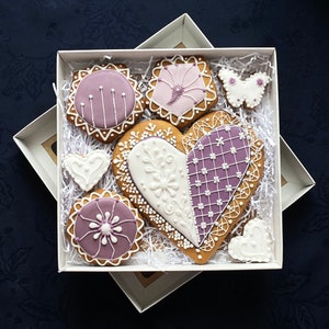 May include: A gift box filled with purple and white decorated gingerbread cookies in the shape of hearts, butterflies, and circles. The cookies are arranged on white shredded paper.