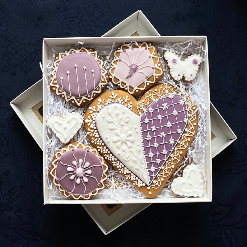 May include: A gift box filled with purple and white decorated gingerbread cookies in the shape of hearts, butterflies, and circles. The cookies are arranged on white shredded paper.