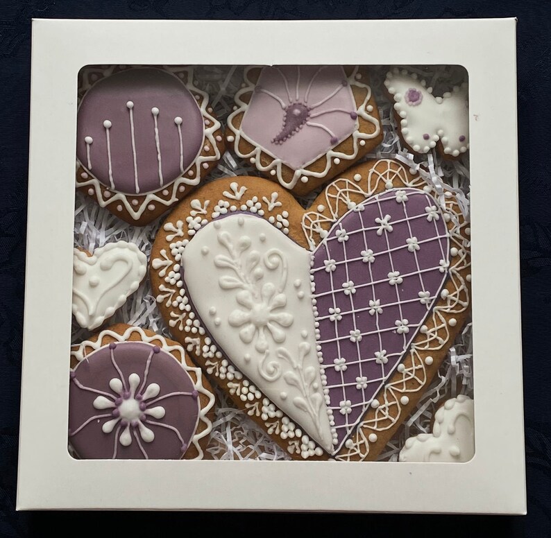 May include: A set of nine purple and white gingerbread cookies in a white box. The cookies are shaped like hearts, circles, and a flower. The cookies are decorated with white icing and purple details.