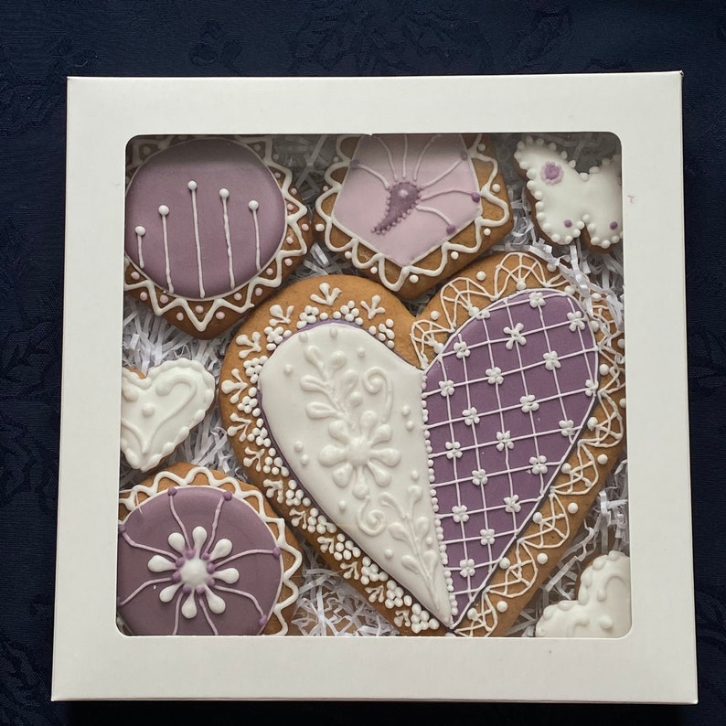 May include: A set of six heart-shaped gingerbread cookies decorated with purple and white icing. The cookies are arranged in a white box with a clear window.