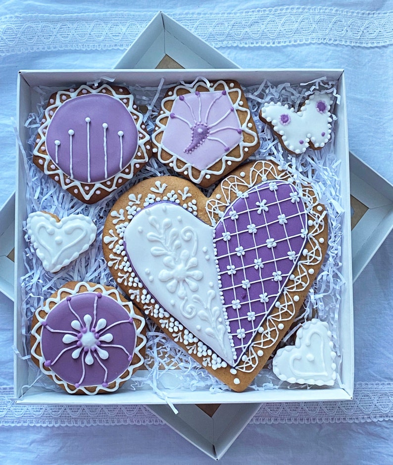 May include: A gift box filled with purple and white decorated gingerbread cookies in the shape of hearts, a butterfly, and a circle. The cookies are arranged on white shredded paper.