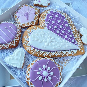 May include: A gift box filled with purple and white decorated gingerbread cookies. The cookies are shaped like hearts, circles, and a butterfly.