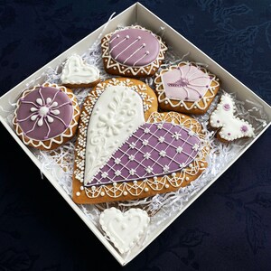 May include: Assortment of purple and white decorated gingerbread cookies in a white gift box. The cookies are shaped like hearts, butterflies, and flowers.
