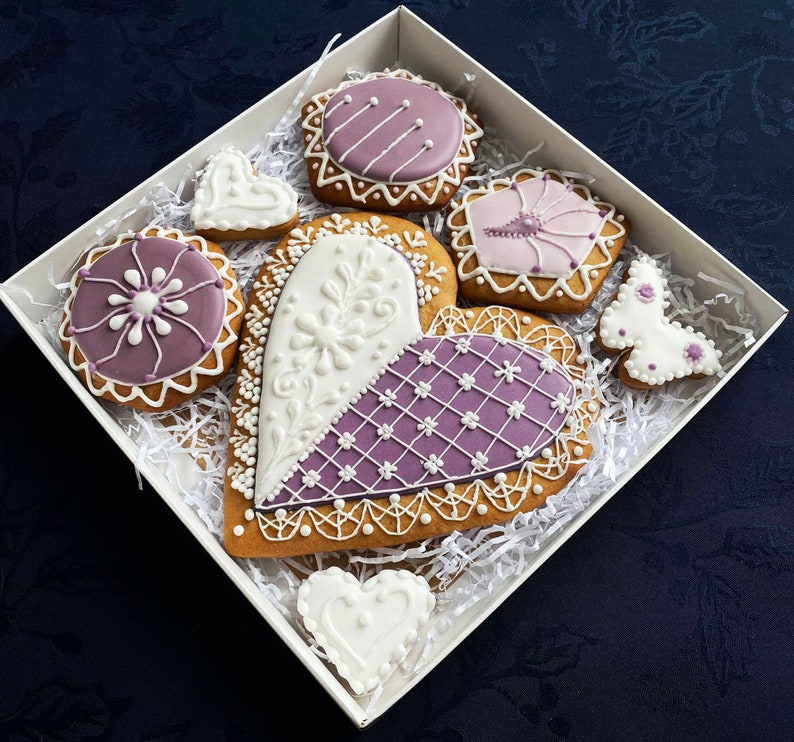 May include: Assortment of purple and white decorated gingerbread cookies in a white gift box. The cookies are shaped like hearts, butterflies, and flowers.